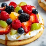 A mini sugar cookie fruit pizza topped with creamy frosting, fresh strawberries, blueberries, raspberries, blackberries, grapes, and a caramel drizzle.