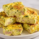 Stacked squares of golden zucchini egg bake with ham and herbs on a white plate.