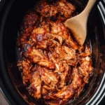 Shredded BBQ chicken in a black crockpot with a wooden spoon, coated in thick, glossy barbecue sauce.