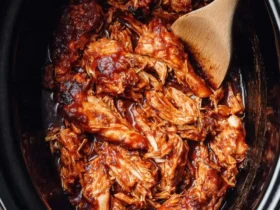 Bbq Chicken Crockpot 44 Shredded BBQ chicken in a black crockpot with a wooden spoon, coated in thick, glossy barbecue sauce.