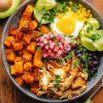 A colorful bbq chicken sweet potato bowl topped with a fried egg, black beans, corn, avocado, and fresh salsa on a wooden table.