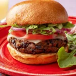 A delicious homemade Black Bean Burger stacked with fresh vegetables and melted cheese on a toasted bun, served on a red plate.