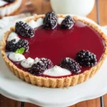 A Blackberry Panna Cotta Tartlet with a graham cracker crust, topped with fresh blackberries, cream dollops, and a glossy berry glaze.