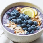 A creamy Blueberry Muffin Smoothie Bowl topped with fresh blueberries, lemon slices, granola, and almond slices in a ceramic bowl.