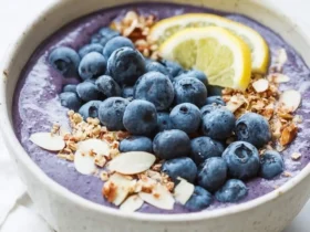 A creamy Blueberry Muffin Smoothie Bowl topped with fresh blueberries, lemon slices, granola, and almond slices in a ceramic bowl.