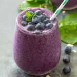 A vibrant purple blueberry spinach smoothie in a glass, garnished with fresh blueberries, a mint leaf, and a green striped straw.