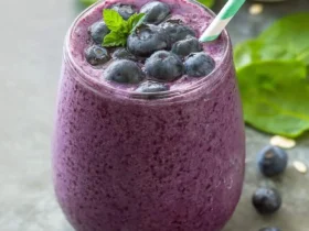 A vibrant purple blueberry spinach smoothie in a glass, garnished with fresh blueberries, a mint leaf, and a green striped straw.