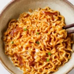 A bowl of creamy Buldak Ramen Carbonara with chopsticks lifting noodles, topped with bacon and green onions.