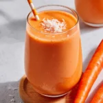 A creamy orange carrot smoothie in a glass topped with coconut flakes and served with a striped straw.