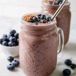 Two mason jars filled with chocolate berry smoothie topped with fresh blueberries and granola, with a metal straw.