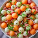 A fresh Grape And Melon Ball Salad with cantaloupe, mozzarella, prosciutto, basil, and red onion in a white bowl.