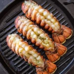 Three Grilled Lobster Tail halves sizzling on a black cast iron grill pan, showing golden-brown shells and tender white meat.