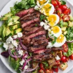 Grilled Steak Cobb Salad with sliced medium-rare steak, hard-boiled eggs, avocado, tomatoes, blue cheese, and red onion on a white plate.