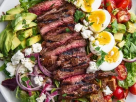 Grilled Steak Cobb Salad with sliced medium-rare steak, hard-boiled eggs, avocado, tomatoes, blue cheese, and red onion on a white plate.
