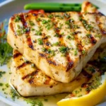 Two perfectly grilled swordfish steaks with char marks, topped with fresh herbs and served with lemon.