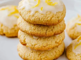 Stack of four glazed Keto Lemon Cookies topped with lemon zest on a white plate with fresh lemons in the background.
