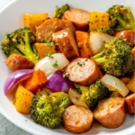 A white bowl filled with One-Pan Honey Garlic Kielbasa And Veggies featuring sliced sausage, broccoli, peppers, and onions.