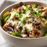 A Philly cheesesteak bowl topped with melted cheese, mushrooms, green peppers, and fresh herbs in a white bowl.
