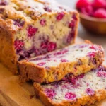 Freshly baked raspberry bread sliced on a wooden cutting board, showing moist texture and vibrant berries inside.