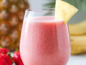 A glass of strawberry banana pineapple smoothie garnished with a pineapple slice on a cork coaster.
