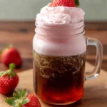 Mason jar with iced coffee topped with strawberry cold foam and fresh strawberry garnish on wooden board.