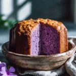 A sliced Ube pound cake with vibrant purple interior sits in a rustic ceramic bowl by a window.