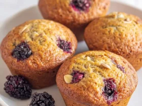 Four golden banana blackberry muffins topped with almond slices sit on a white plate with fresh blackberries.