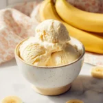 A ceramic bowl filled with creamy banana ice cream, surrounded by fresh banana slices and whole bananas in the background.
