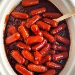 Bbq Smokies Crockpot filled with glossy mini sausages coated in rich barbecue sauce and a wooden spoon.