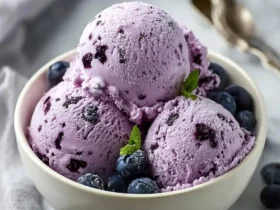 Three scoops of creamy blueberry ice cream with cottage cheese in a white bowl, topped with fresh blueberries and mint leaves.
