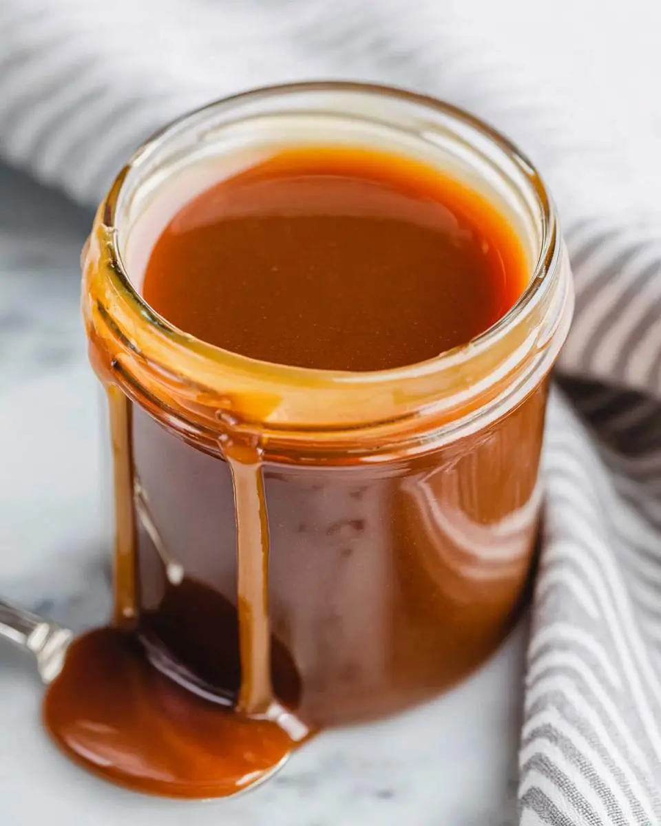 A glass jar filled with rich, glossy caramel sauce with drips running down the side onto a marble surface.