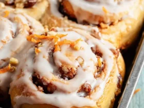 Freshly baked Carrot Cake Cinnamon Rolls topped with cream cheese glaze, chopped walnuts, and orange zest in a baking pan.