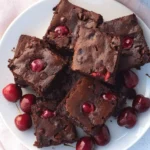 Stack of fudgy Chocolate Cherry Brownies with fresh cherries and chocolate chips on a white plate.