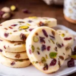 Stack of round Cranberry Pistachio Shortbread cookies on a white plate, showing colorful dried cranberries and green pistachio pieces throughout the buttery dough.