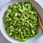 Cucumber Edamame Salad with sesame seeds and fresh herbs served in a white speckled bowl with a wooden spoon.