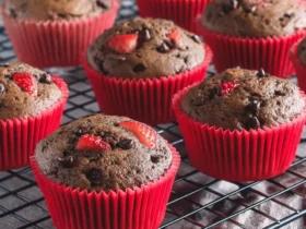 Double Chocolate Strawberry Muffins 47 Freshly baked Double Chocolate Strawberry Muffins in red liners cooling on a wire rack with strawberries in background.
