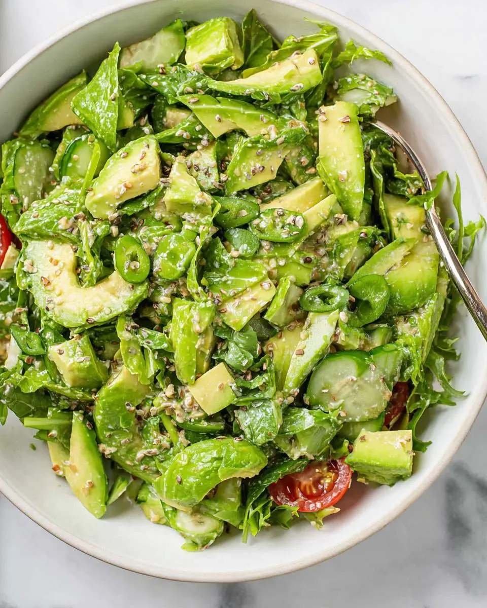 Fresh Green Goddess Avocado Salad with cucumber, spinach, and sesame seeds in a white bowl with a serving spoon.