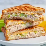 Three halves of a grilled tuna sandwich stacked on a white plate, showing melted cheese and celery inside.