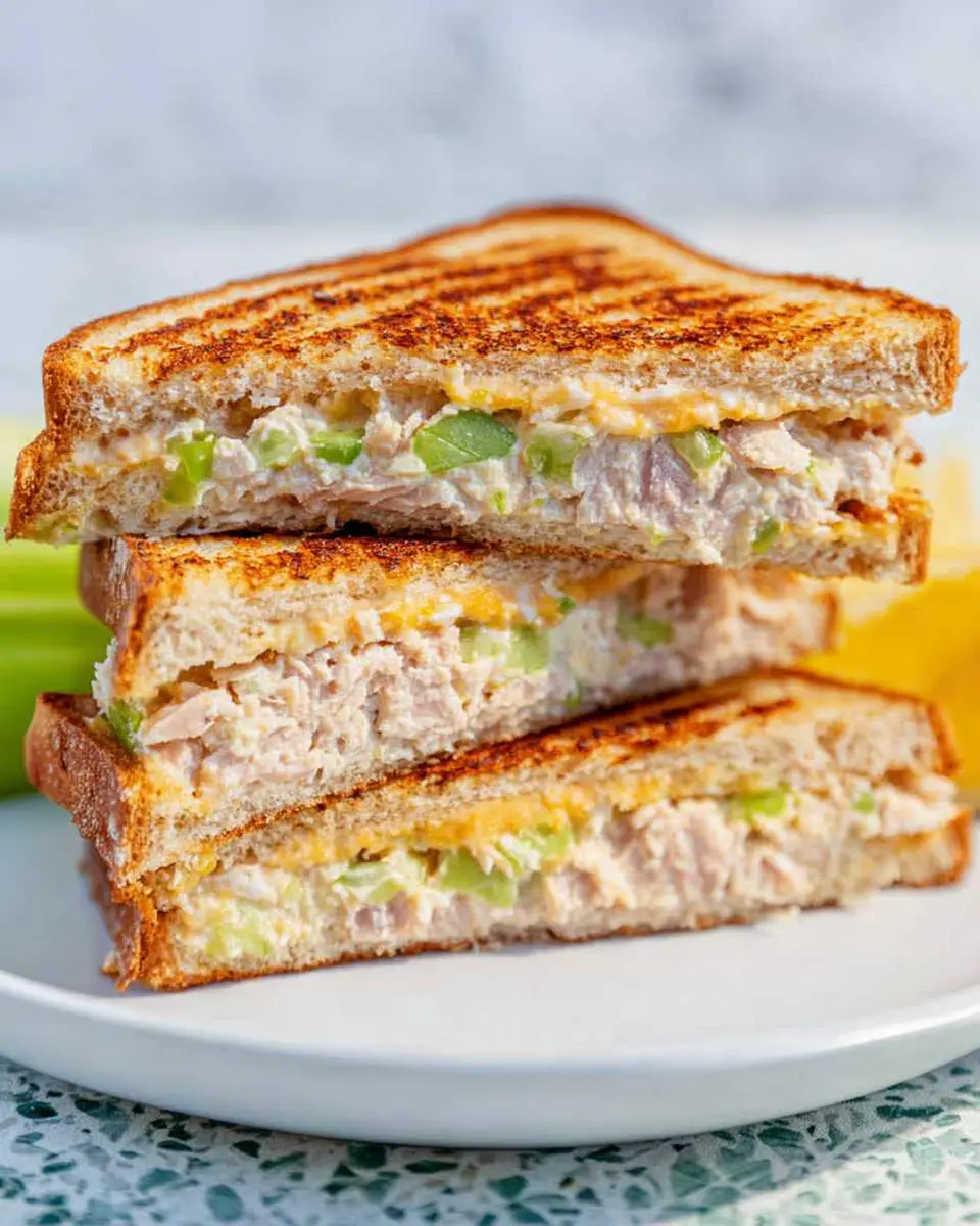 Three halves of a grilled tuna sandwich stacked on a white plate, showing melted cheese and celery inside.