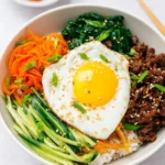 Korean Bibimbap Beef Bowl with rice, seasoned beef, vegetables, and a fried egg topped with sesame seeds.