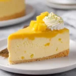 A slice of Mango Mousse Cake topped with fresh mango chunks, whipped cream, and lime zest on a white plate.