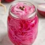 A glass mason jar filled with vibrant pink Pickled Red Onions topped with black peppercorns.