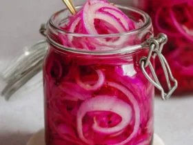Glass jar filled with vibrant Pickled Red Onions, with a golden spoon lifting sliced rings from the brine.
