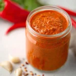 A glass jar filled with vibrant Red Pepper Pesto sits on a marble surface with fresh peppers and garlic nearby.