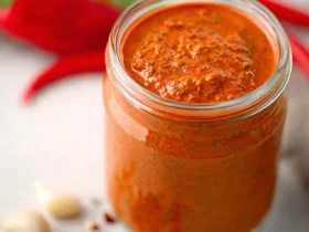 Red Pepper Pesto 37 A glass jar filled with vibrant Red Pepper Pesto sits on a marble surface with fresh peppers and garlic nearby.