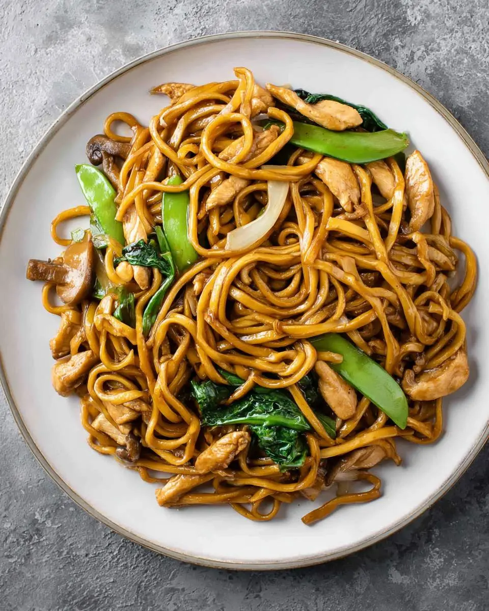 Plate of Shanghai Stir-Fried Noodles With Chicken featuring thick noodles, tender chicken pieces, snow peas, mushrooms, and leafy greens.