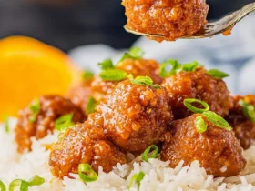 A fork lifts a glazed Slow Cooker Orange Meatball above a plate of white rice topped with more meatballs and green onions.