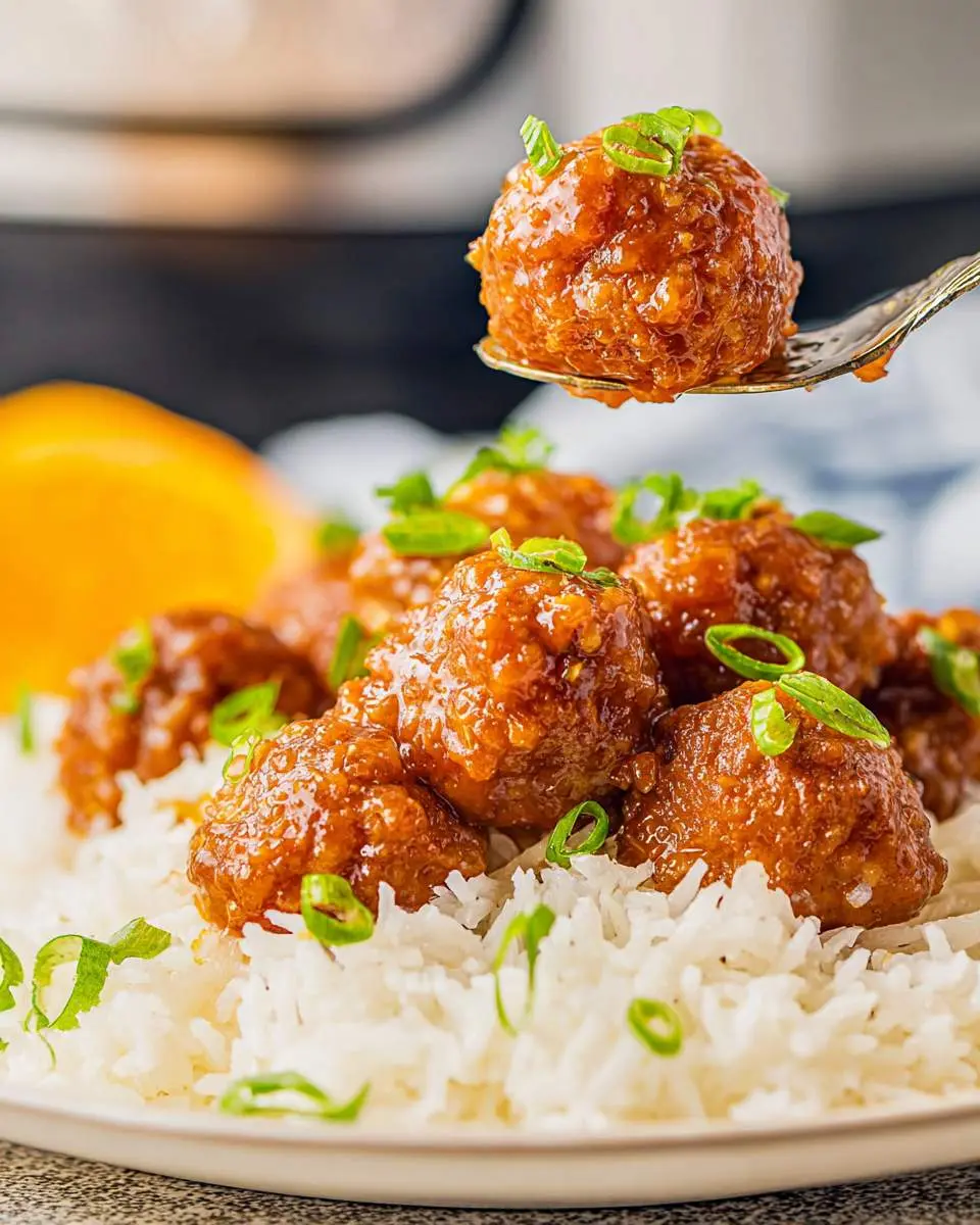 Slow Cooker Orange Meatballs 28 A fork lifts a glazed Slow Cooker Orange Meatball above a plate of white rice topped with more meatballs and green onions.