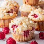 Freshly baked White Chocolate Raspberry Muffins topped with white chocolate chips, surrounded by fresh raspberries on a marble surface.