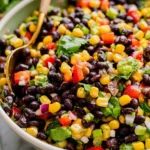 Close-up of a colorful black bean salad with corn, tomatoes, cilantro, and red onion in a white bowl with a gold spoon.